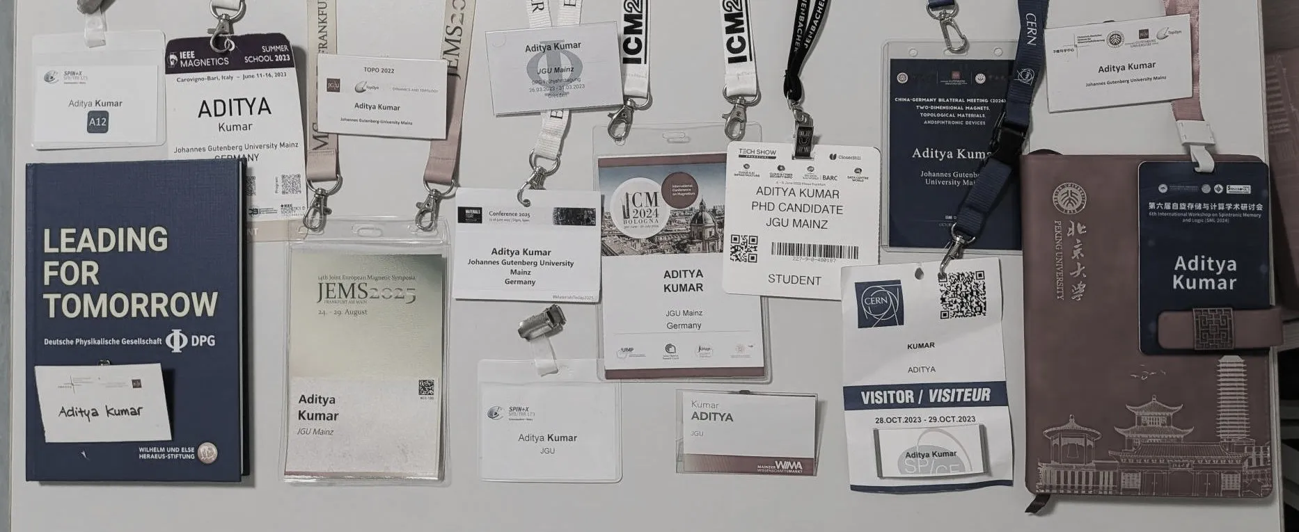 A dozen conference name badges bearing Aditya Kumar and Johannes Gutenberg University Mainz, laid out with their lanyards on a table, among them ICM 2024 Bologna, JEMS 2025 Frankfurt, the Materials Today Conference 2025, the 2024 China-Germany bilateral meeting, an IEEE Magnetics summer school, a CERN visitor pass and a Peking University notebook, alongside a blue DPG "Leading for Tomorrow" volume.