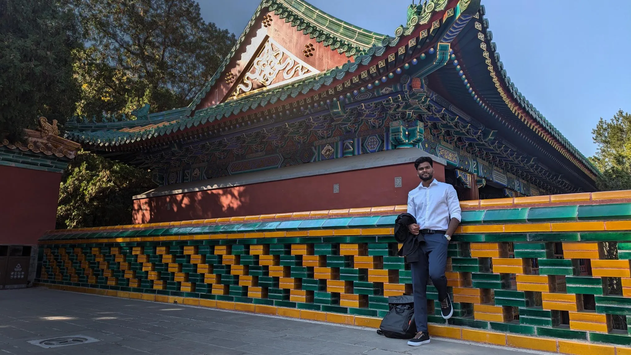 Aditya Kumar leaning against the glazed green and yellow tiled balustrade of a Chinese temple courtyard in Beijing, in a white shirt with a jacket over his arm, the painted eaves of the temple rising behind him.