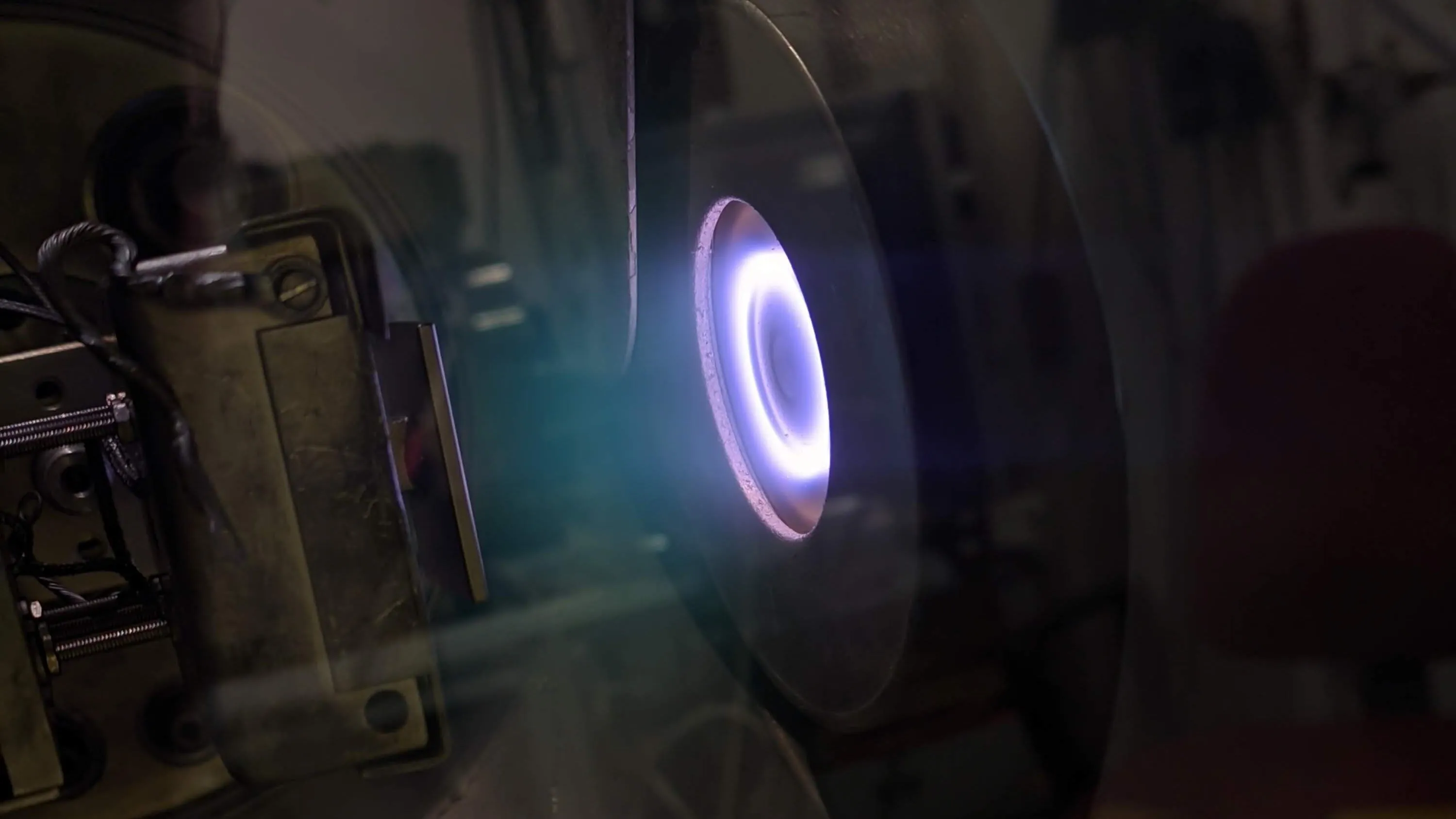 A sputter deposition source seen through the port of a darkened vacuum chamber, its target ringed by a bright violet-white plasma glow.