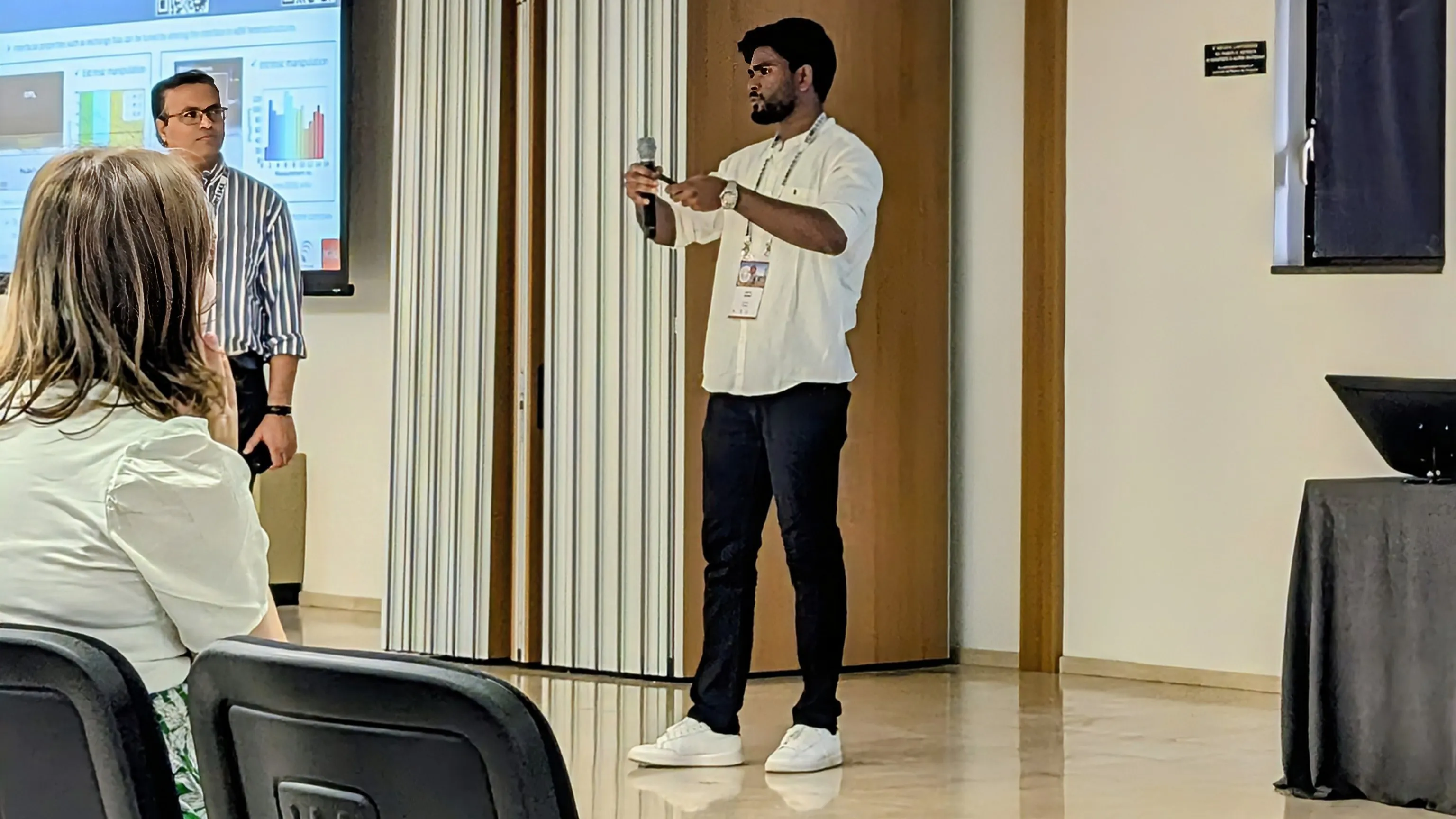 Aditya Kumar speaking at the ICM 2024 conference in Bologna: standing with a microphone in a white shirt and conference lanyard, a co-presenter beside a projected slide of bar charts behind him, and the seated audience in the foreground.