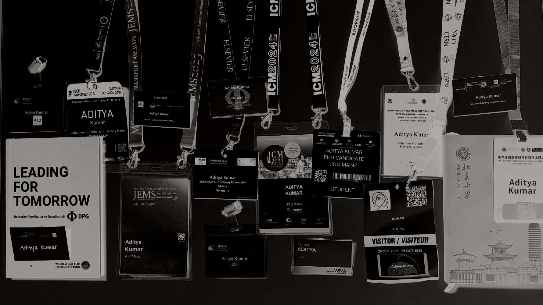 A dozen conference name badges bearing Aditya Kumar and Johannes Gutenberg University Mainz, laid out with their lanyards, shown as a black-and-white negative on a black ground, among them ICM 2024 Bologna, JEMS 2025 Frankfurt, the Materials Today Conference 2025, the 2024 China-Germany bilateral meeting, an IEEE Magnetics summer school, a CERN visitor pass and a Peking University notebook, alongside a DPG "Leading for Tomorrow" volume.
