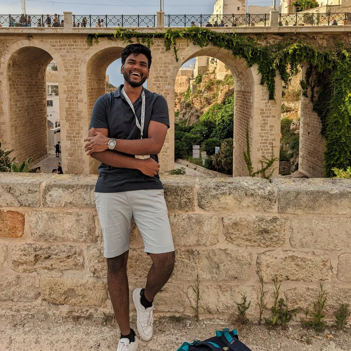 Aditya Kumar leaning against a low stone wall in Polignano a Mare, laughing, wearing a conference lanyard, with the town's arched bridge and the sea gorge behind him.