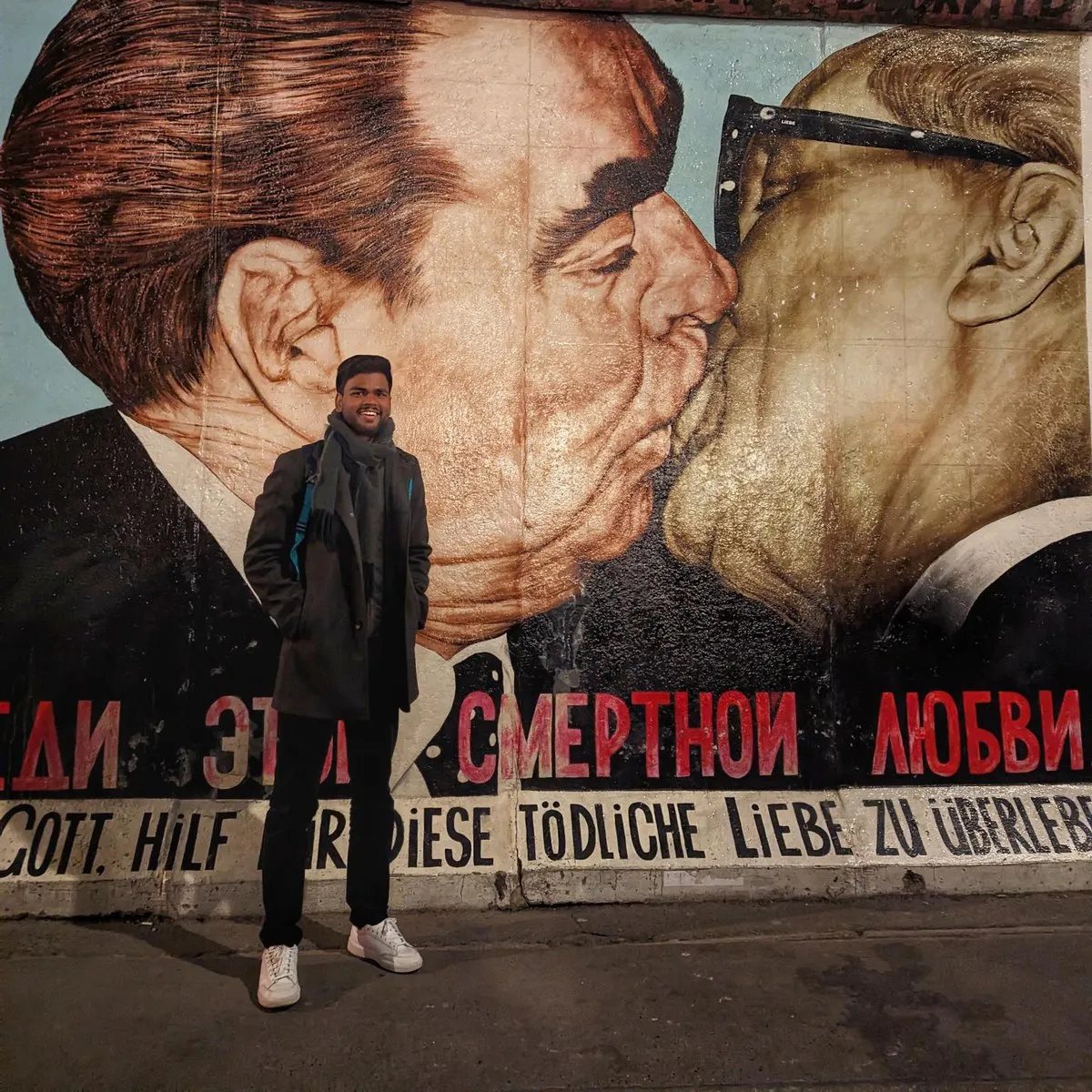 Aditya Kumar standing at night in front of the East Side Gallery section of the Berlin Wall, below the mural of Brezhnev and Honecker kissing.