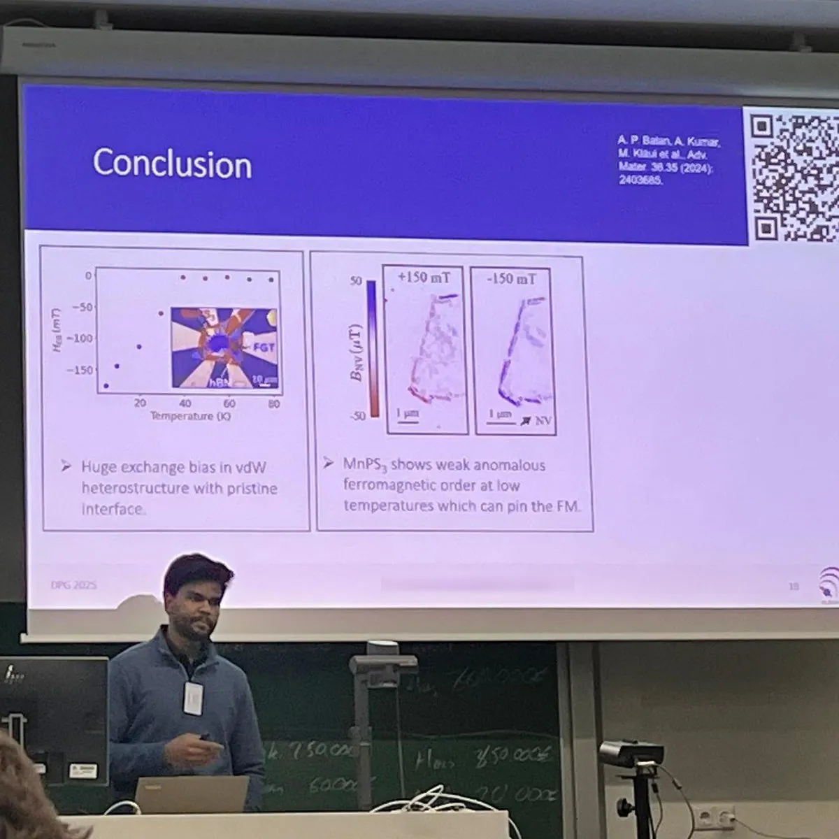Aditya Kumar speaking at a lectern in a lecture hall, below a projected slide headed Conclusion showing an exchange-bias field against temperature and two magnetic images of a flake at plus and minus 150 millitesla.