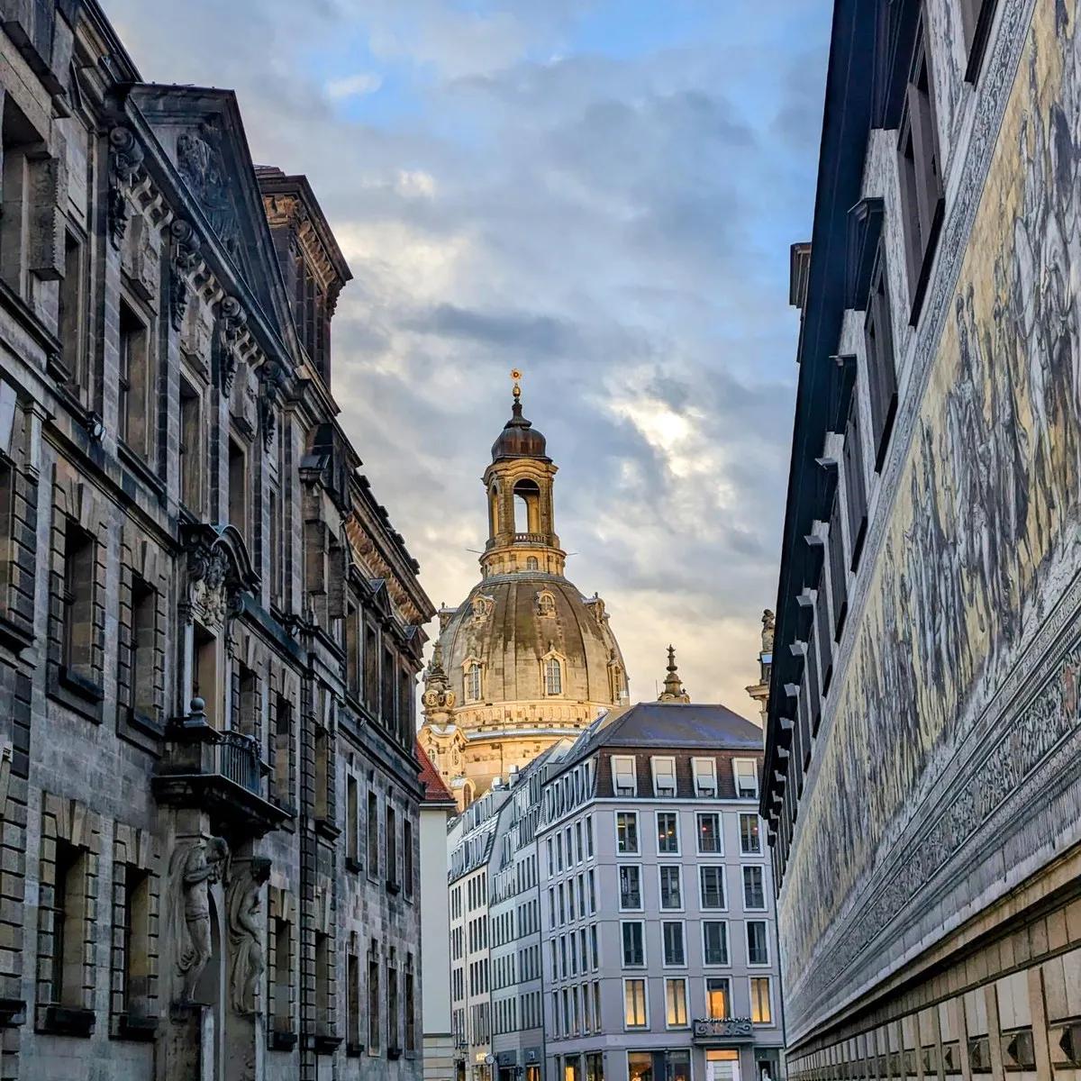The dome of the Dresden Frauenkirche at dusk, seen down a narrow street between the Fürstenzug mural on the right and a baroque facade on the left.