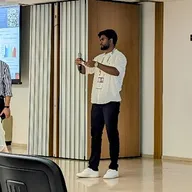 Aditya Kumar speaking into a handheld microphone during his invited talk at ICM 2024 in Bologna, with a colleague and a projected slide visible behind him.