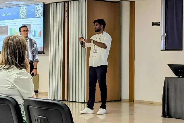 Aditya Kumar speaking into a handheld microphone during his invited talk at ICM 2024 in Bologna, with a colleague and a projected slide visible behind him.