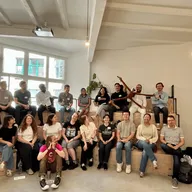 About two dozen participants of the DPG Leading for Tomorrow 2026 programme seated in two rows on stepped wooden benches in a bright room, most wearing name badges, one holding a large stuffed gibbon with outstretched arms, with a yellow-green DPG banner standing at the right.