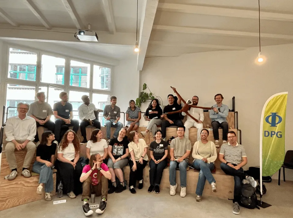 About two dozen participants of the DPG Leading for Tomorrow 2026 programme seated in two rows on stepped wooden benches in a bright room, most wearing name badges, one holding a large stuffed gibbon with outstretched arms, with a yellow-green DPG banner standing at the right.