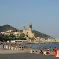 The seafront at Sitges, Spain: a sandy beach with sunbathers, swimmers and rows of closed white parasols, backed by a palm-lined promenade of white buildings, with a large stone church and its bell tower standing on the promontory beyond and a wooded green hill rising behind it under a clear blue sky.